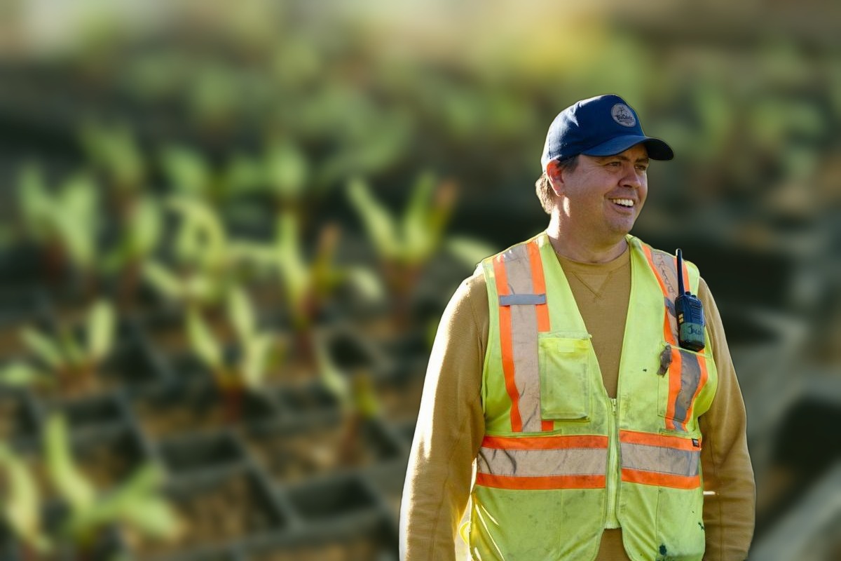 Jacob Beaton smiling in a high-visibility vest and ball cap, with seedlings growing in trays behind him.